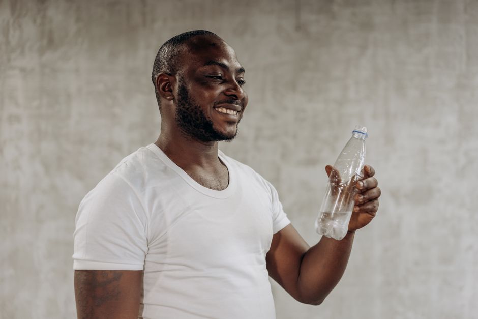 A happy man smiles while holding a bottle of water indoors, embracing healthy living.