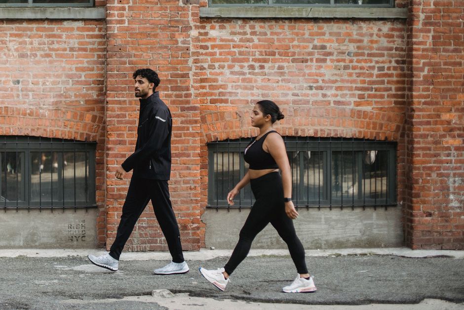 A man and woman walking briskly in sportswear against a brick wall in the city.