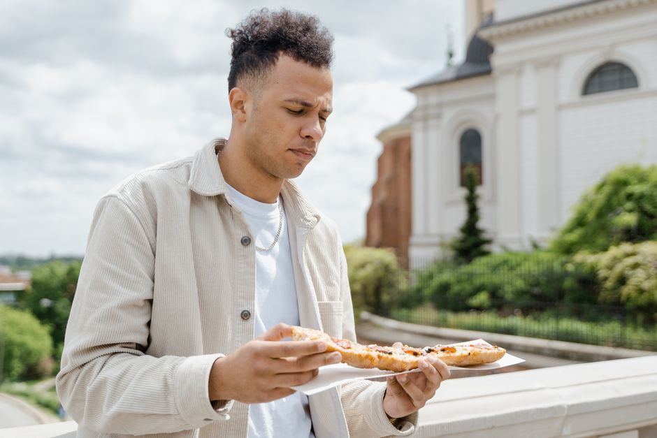 A man in a beige jacket enjoys a slice of pizza in front of a historic building on a sunny day.