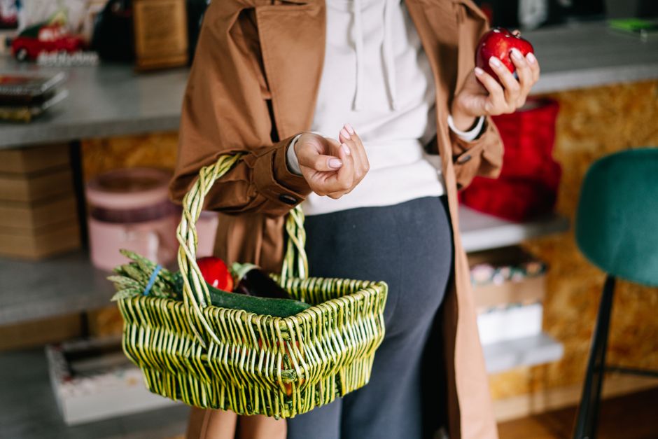 A pregnant woman holds a green wicker basket filled with fresh apples indoors.