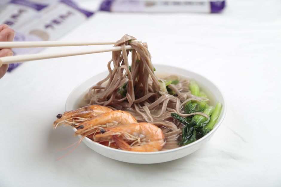 A steaming bowl of soba noodles with shrimp and greens, elegantly lifted by chopsticks.