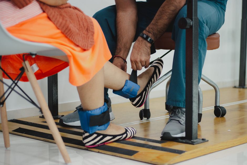 A therapist assists a woman during a rehabilitation session involving ankle weights.