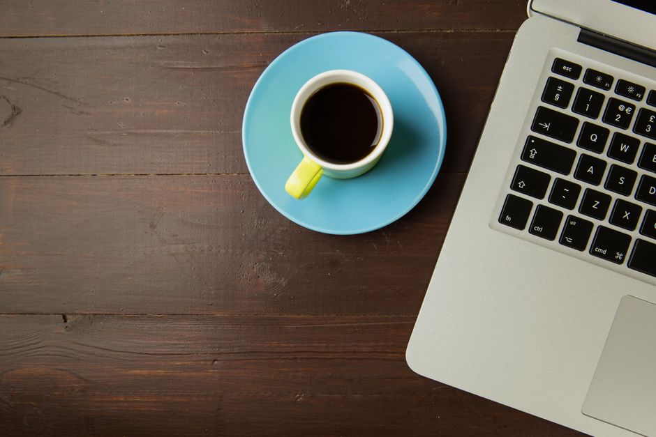 A top view of a coffee cup on a saucer next to a laptop on a wooden desk, ideal for work-from-home themes.