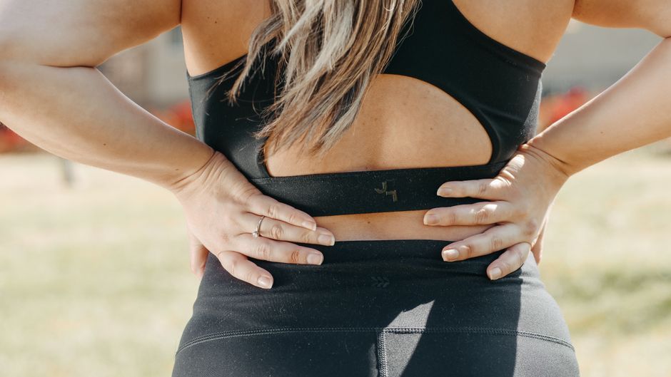 A woman in activewear holds her lower back, indicating muscle strain, outdoors.