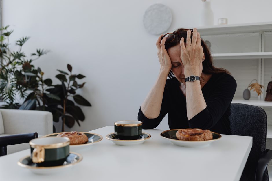 A woman in emotional distress sits at a table with coffee and pastries, illustrating loneliness and sadness.