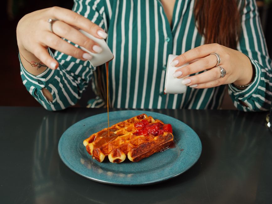 A woman pours syrup over a waffle topped with jam on a blue plate, showcasing breakfast indulgence.