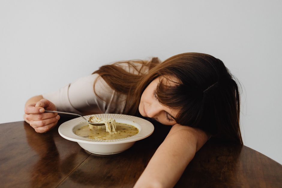 A woman resting her head on the table while holding a spoon and eating soup, conveying fatigue.