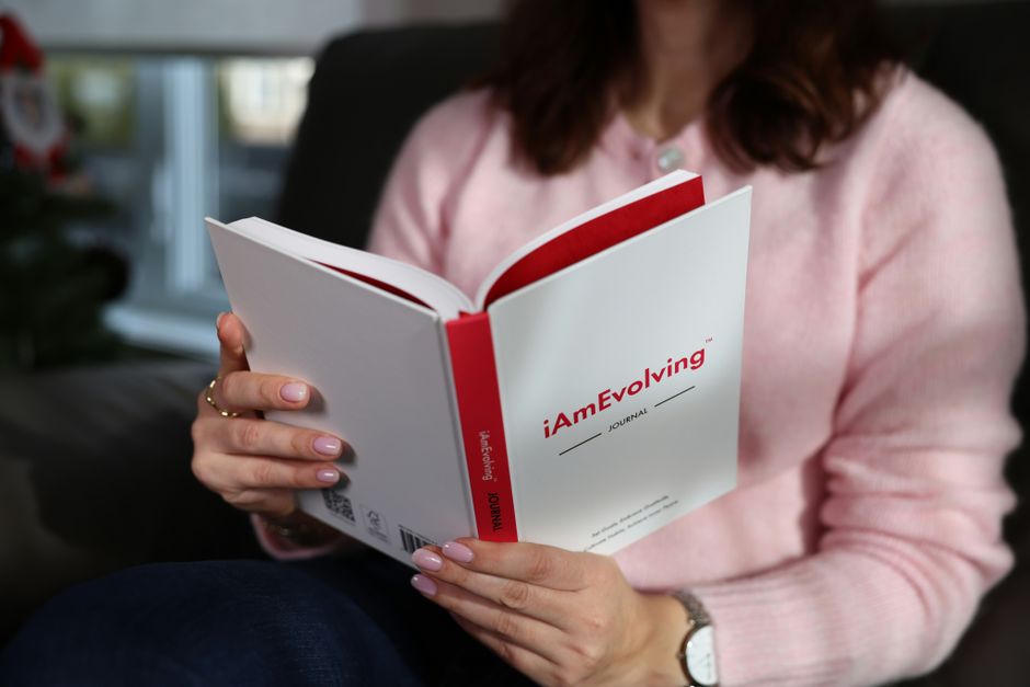 A woman with pink nails and sweater reads a 'iAmEvolving' journal, promoting mindfulness.