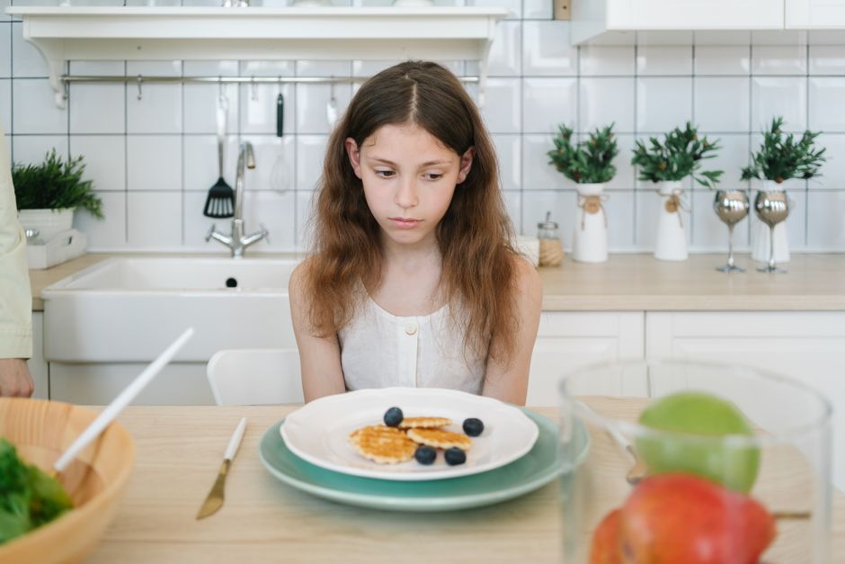A young girl looks down unhappily at her breakfast pancakes with blueberries in a modern kitchen.