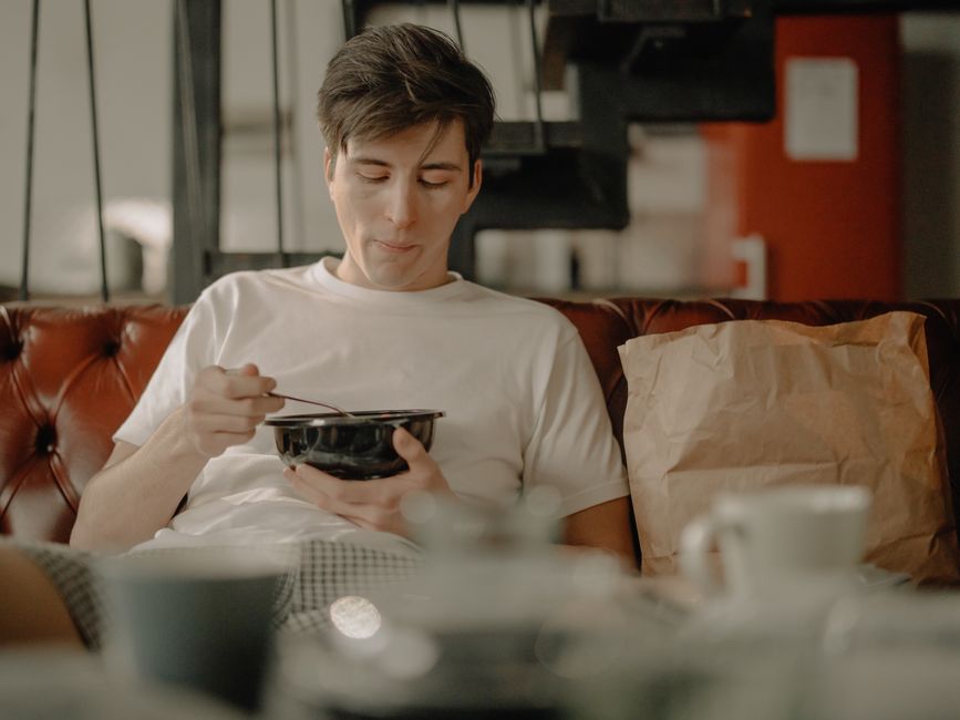A young man relaxes at home, enjoying a meal with a takeout bag on the couch.