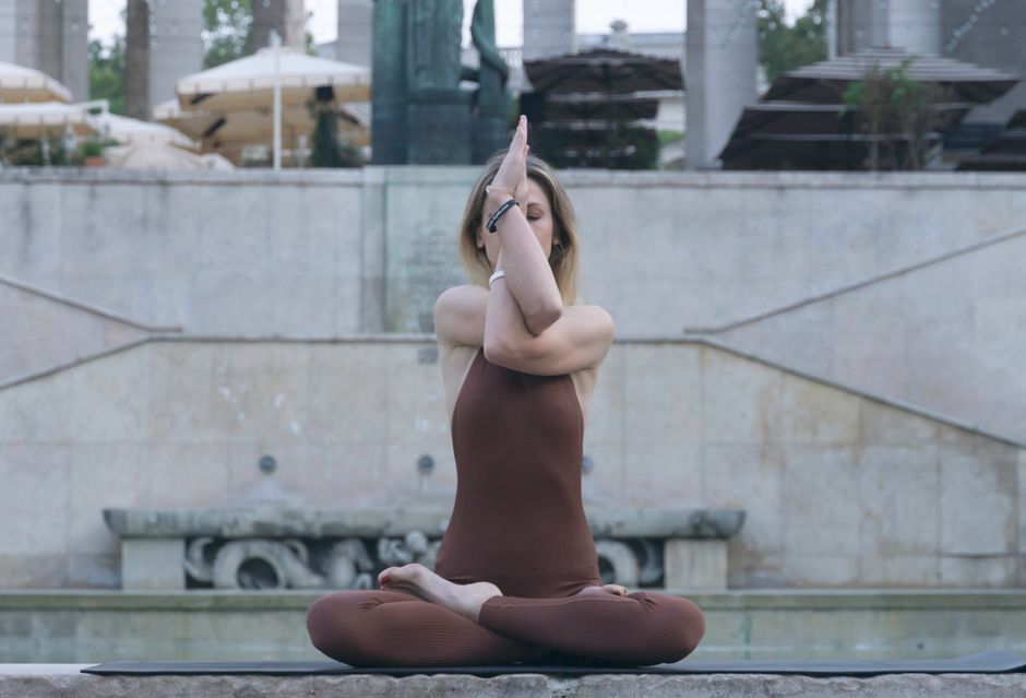 A young woman meditates in a yoga pose by an outdoor fountain, promoting relaxation.
