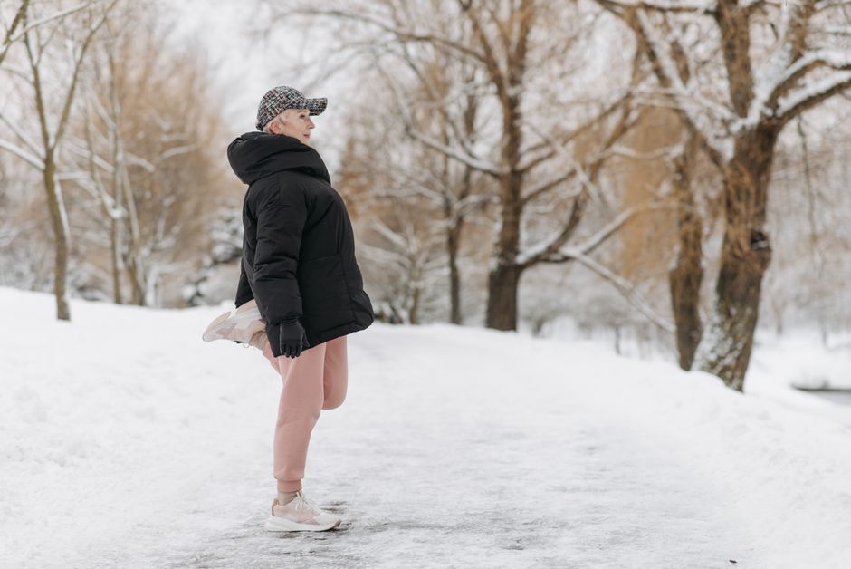 An elderly woman warms up for exercise in a snowy park.