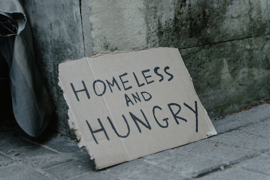 Cardboard sign with 'Homeless and Hungry' message leaning against a wall on a sidewalk.
