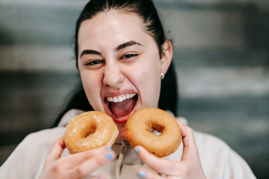 Cheerful young female with dark hair and opened mouth holding tasty doughnuts and eating dessert despite diet