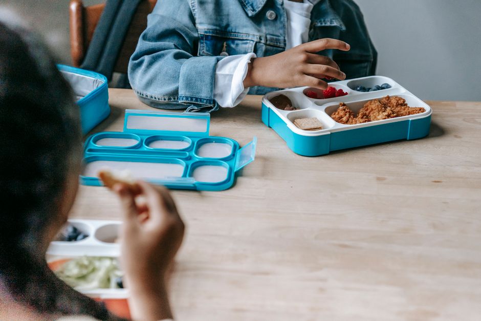 Crop faceless classmates sitting at table with lunch boxes with various food and eating during break