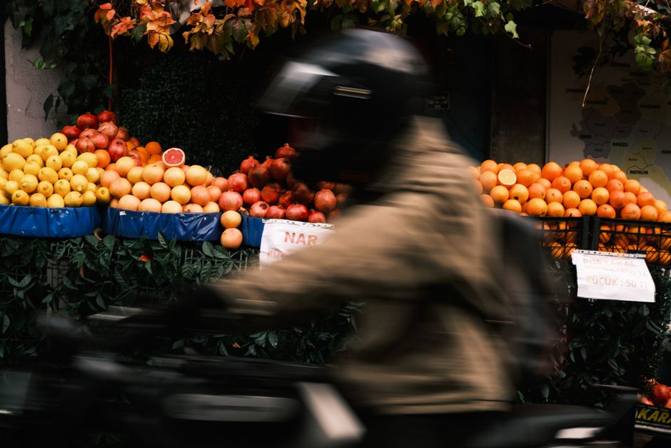 Dynamic shot of a motorcyclist speeding past a vibrant fruit stand with apples and oranges.