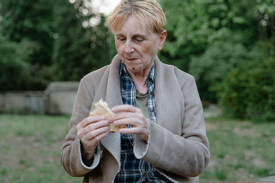 Elderly woman with short hair eating a sandwich in a serene outdoor setting.