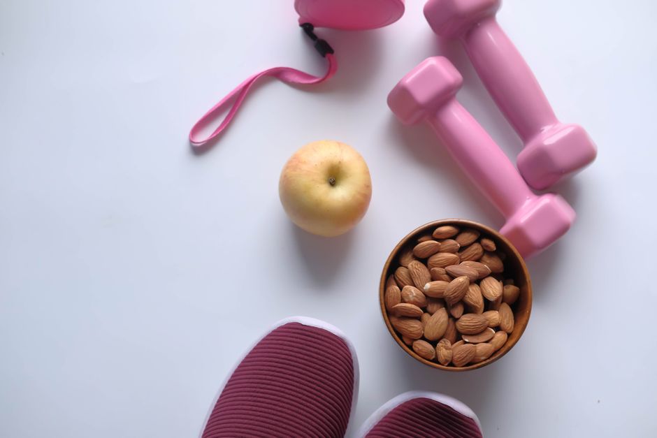 Flat lay of fitness essentials: pink dumbbells, almonds, apple, and sneakers on a white background.