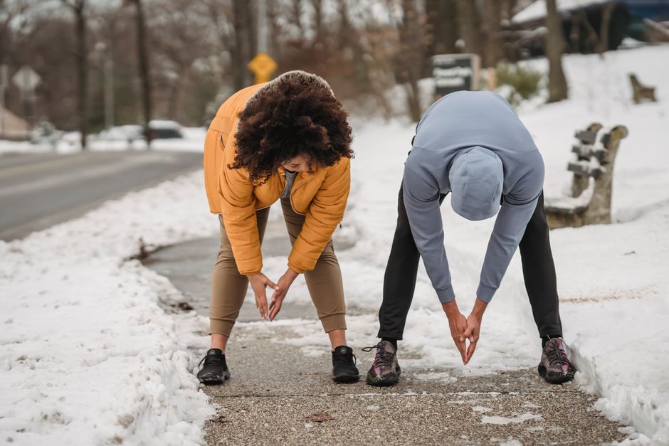 Full length faceless ethnic male trainer and plump female stretching body and bending down before working out on snowy sidewalk on winter day