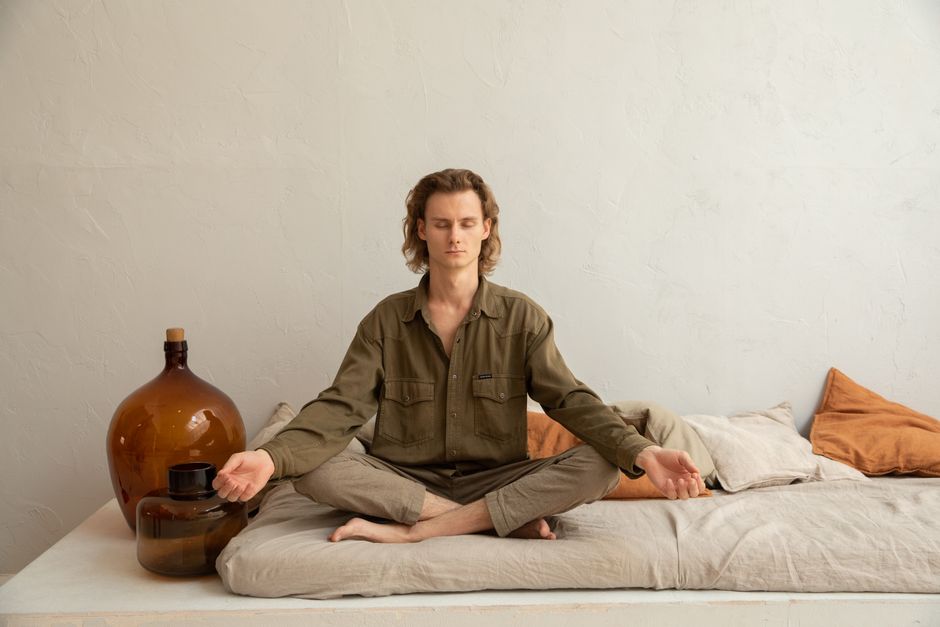 Full length young relaxed male in casual wear sitting with eyes closed in Lotus Pose on soft mattress and meditating