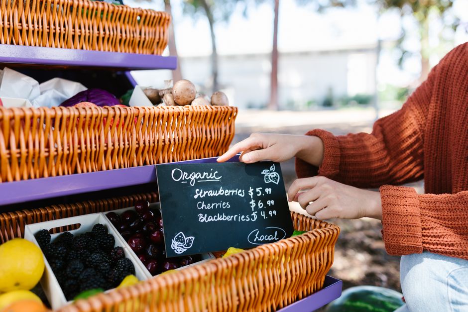 Hands arranging organic fruits and a blackboard at an outdoor farmer's market stand.