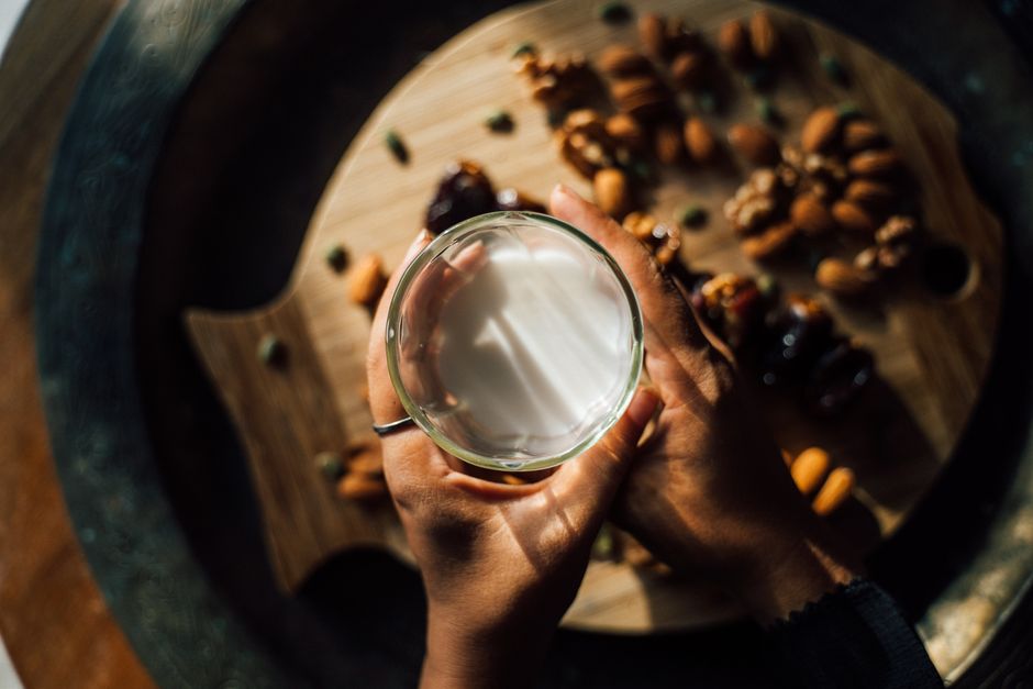Hands holding a glass of milk above nuts on a wooden board, captured from above.
