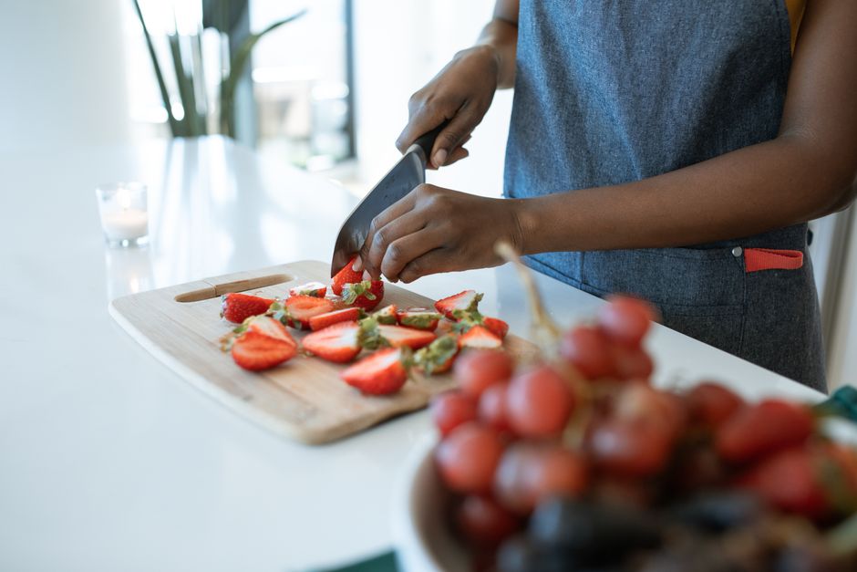 Hands slicing strawberries on a chopping board, with grapes in the foreground.
