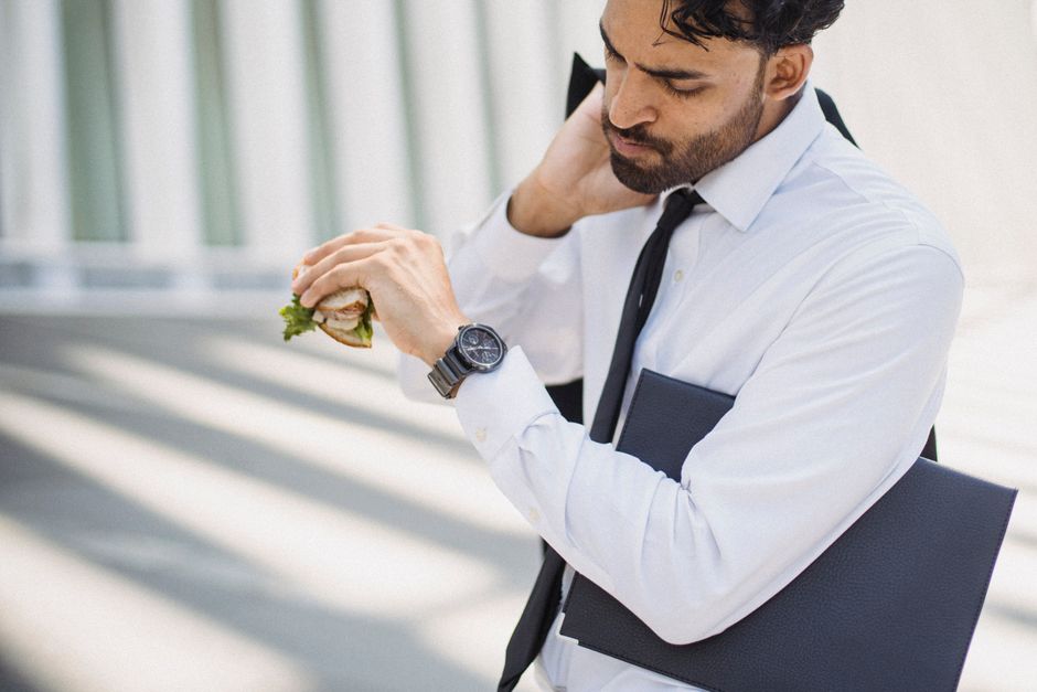 Professional man in corporate attire checking time while holding a sandwich outdoors.