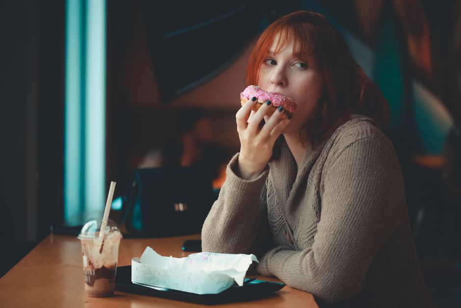 Red-haired woman savoring a pink doughnut in an indoor cafe setting. Warm, inviting ambiance.