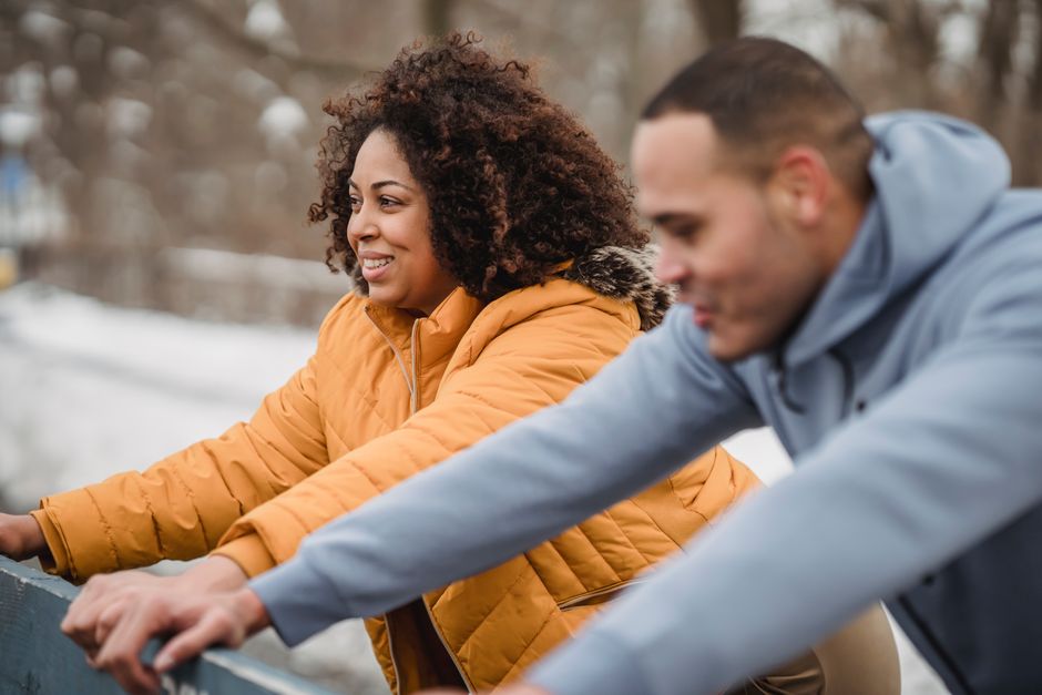 Side view of positive African American couple leaning on structure while doing warm up exercises on snowy street on blurred background