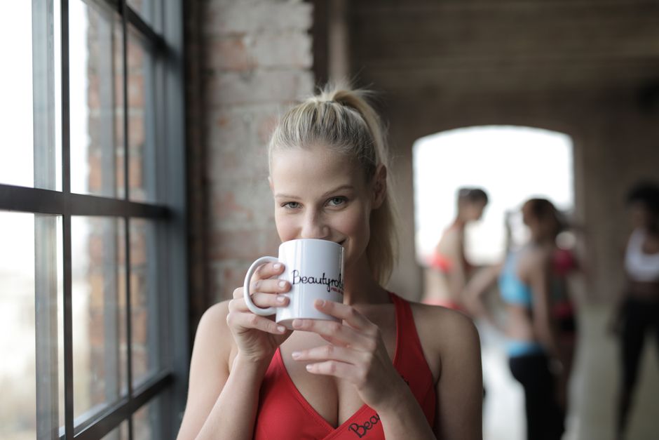 Smiling young blond female in sportswear looking at camera while standing by window in gym and drinking tea