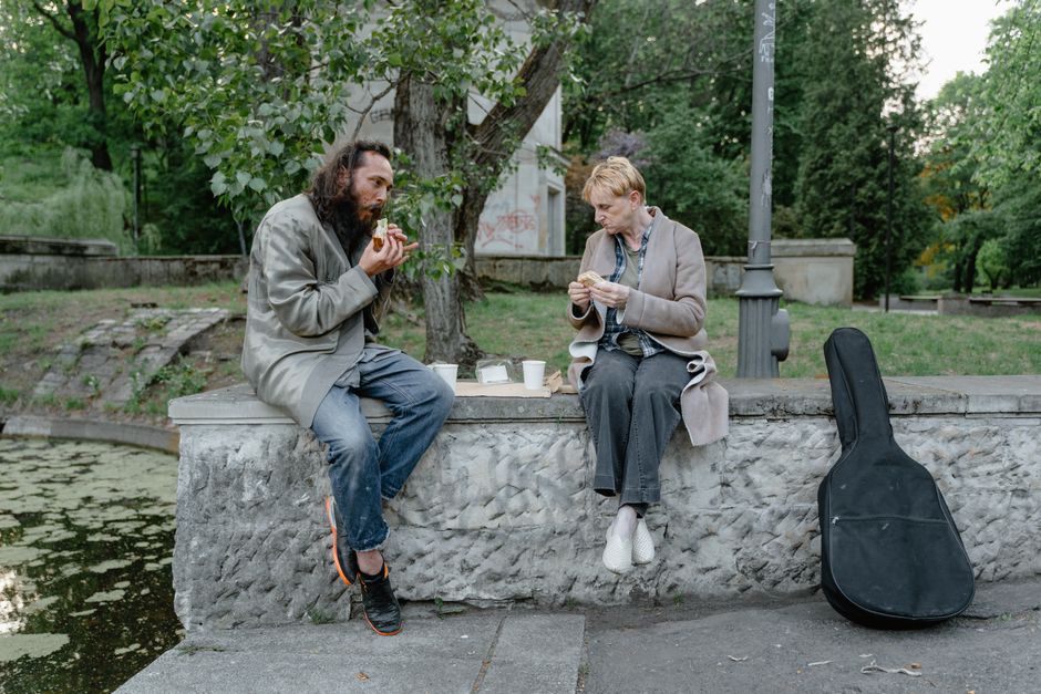Two individuals sharing a meal outdoors on a stone bench, highlighting companionship and resilience.