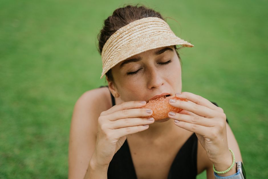 Woman savoring a snack outdoors with eyes closed, wearing a sun visor for relaxation.