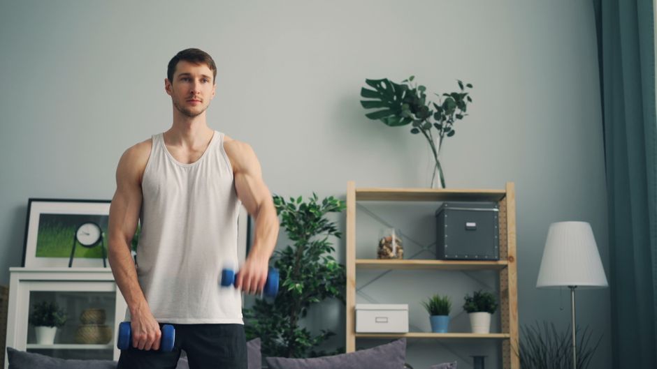 A muscular man in a tank top exercises with dumbbells indoors during the day.