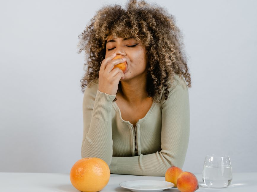A woman savoring a fresh peach at a table with fruit, embodying healthy eating.