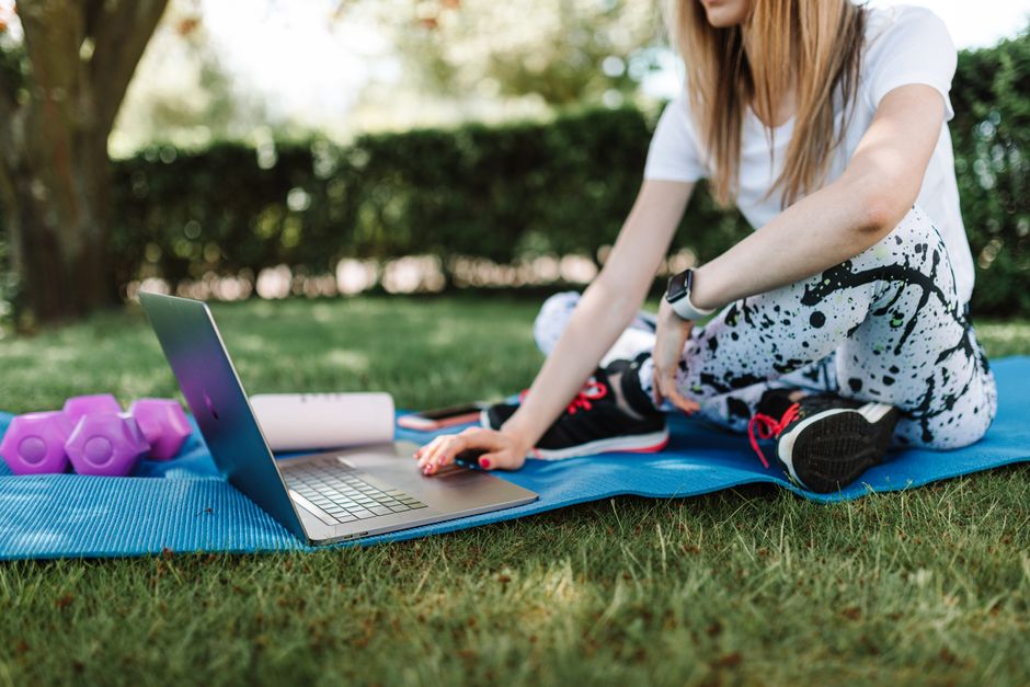 A woman sits on a yoga mat outdoors using a laptop for fitness guidance.
