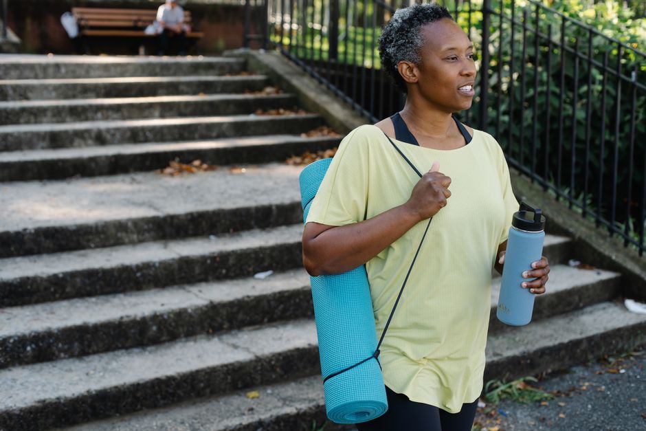 African American woman walking outdoors with yoga mat and tumbler promoting healthy lifestyle.