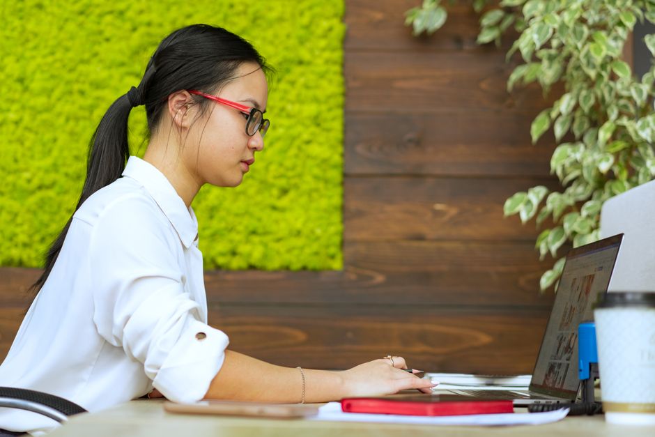 Asian woman working remotely on laptop in a stylish, modern office setting with green decor.