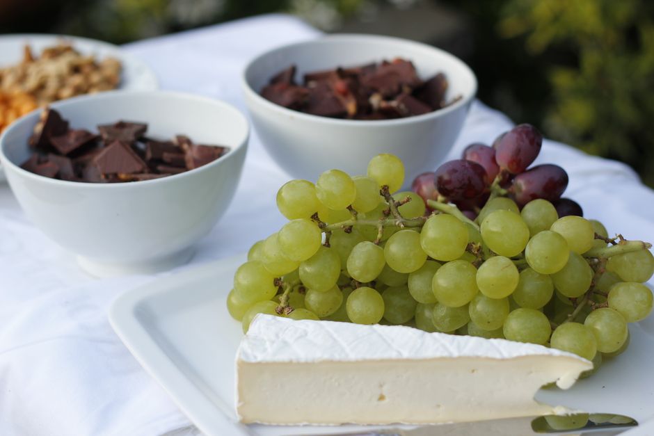 Close-up of a cheese platter with grapes, chocolate, and nuts on a sunny white table setting.