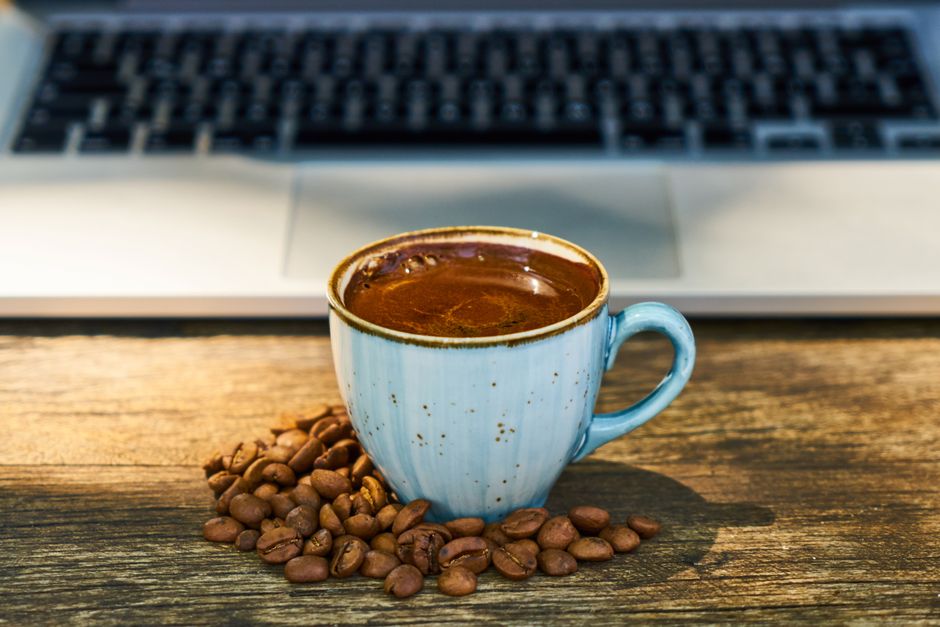 Close-up of Turkish coffee cup surrounded by beans near a laptop, wooden background.
