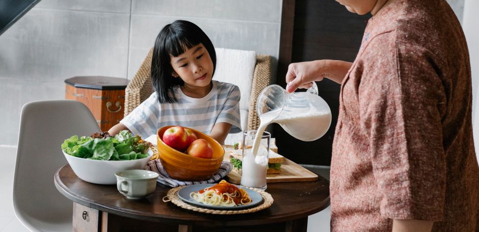 Crop faceless female standing near table and preparing healthy lunch for cute Asian kid at home