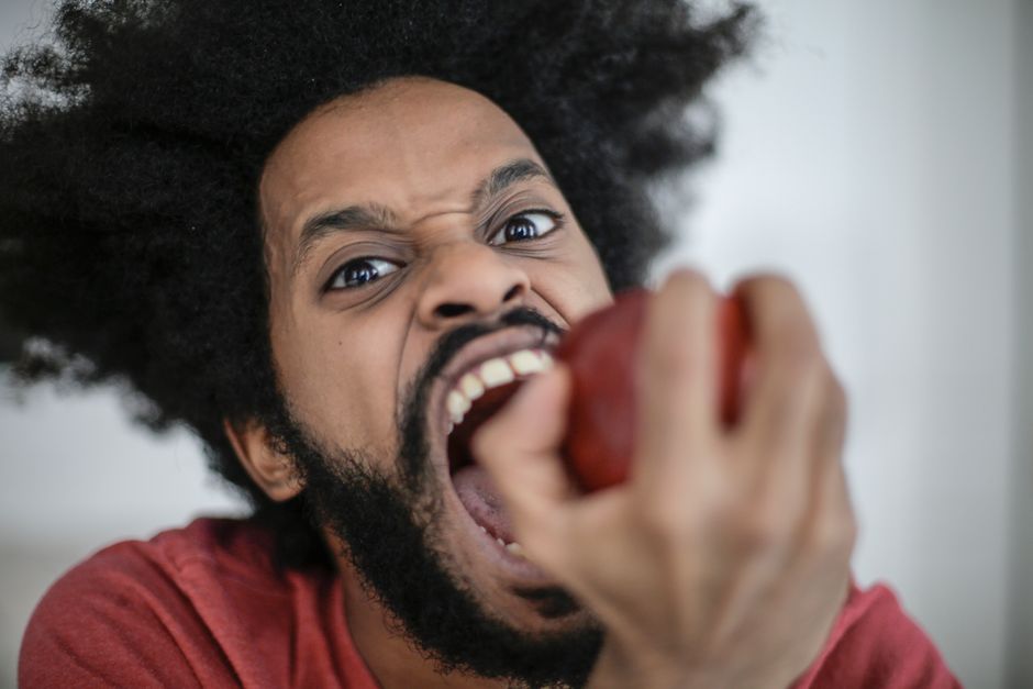 Energetic portrait of a man with afro hair biting into a red apple with an intense expression.