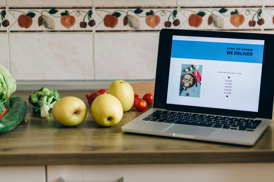 Laptop showing a delivery app beside fresh fruits and vegetables on a kitchen counter.