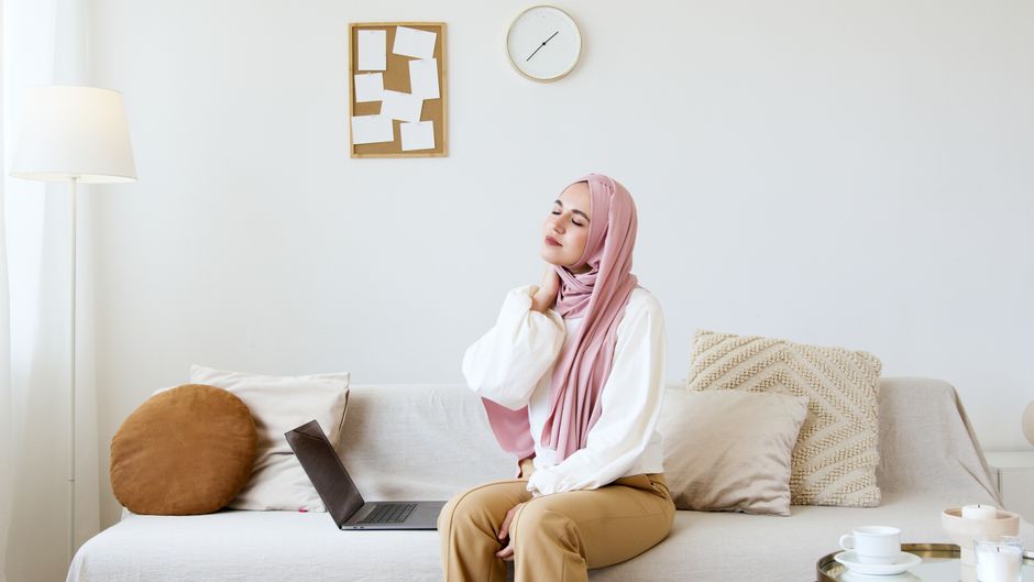 Muslim woman in hijab sitting on sofa, relaxing with eyes closed and hand on neck. Modern living room setting with laptop.