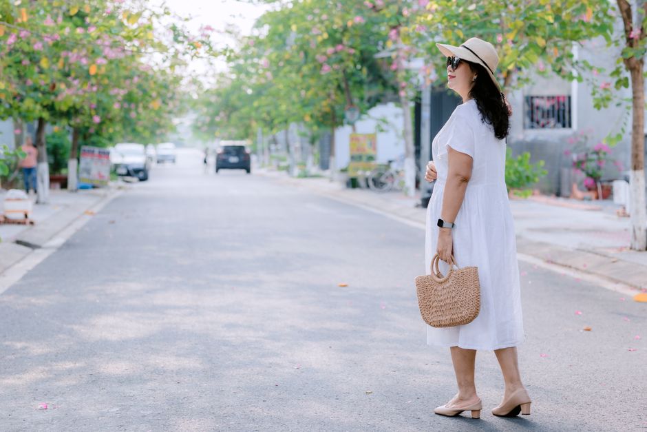 Pregnant woman in white dress and sunhat walking on a sunny, tree-lined street.