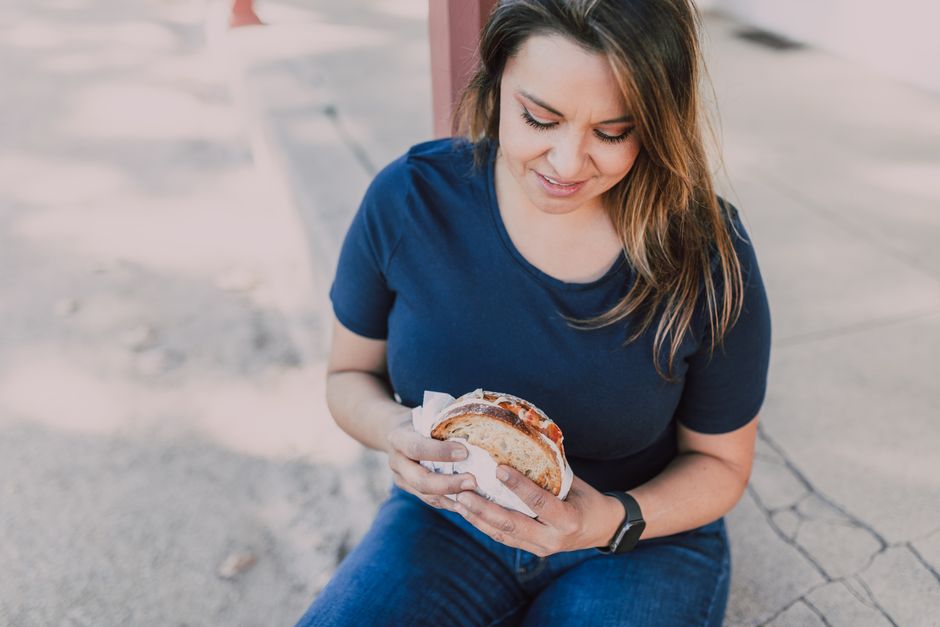 Smiling woman holding a sandwich on a sunny day, enjoying a casual outdoor meal.