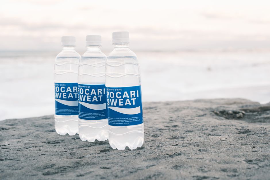 Three Pocari Sweat bottles placed on a beach rock by the sea.