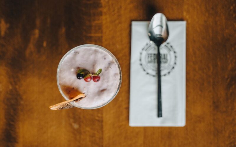 Top view of a creamy berry smoothie garnished with fresh herbs and a biscuit, beside a neatly placed spoon.