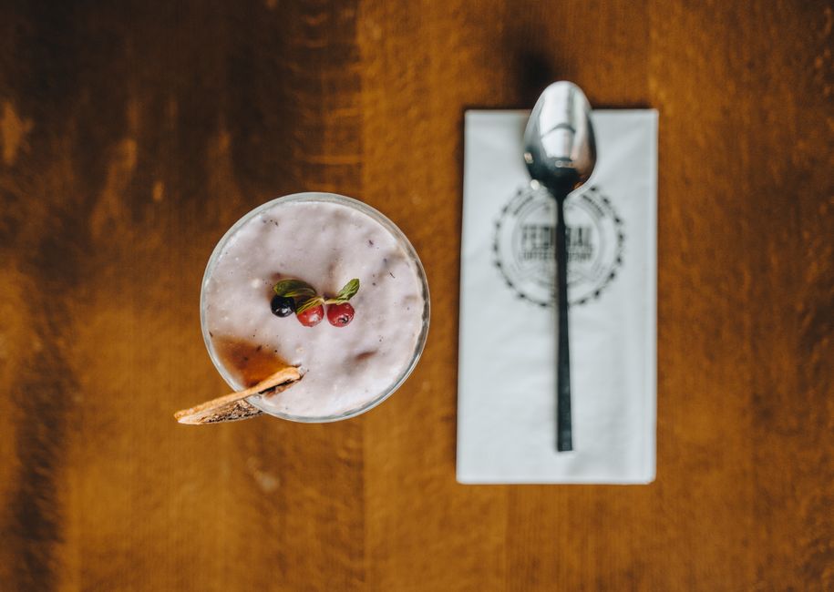 Top view of a creamy berry smoothie garnished with fresh herbs and a biscuit, beside a neatly placed spoon.