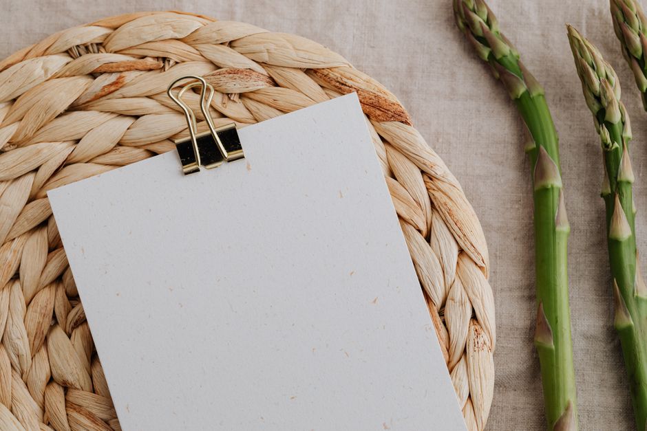 Top view of fresh asparagus next to a blank paper with a clip on rustic woven mat.